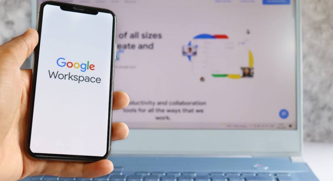 Hand holding a phone displaying Google Workspace logo in front of a laptop also displaying Google Workspace.
