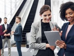 A young woman in business attire stands with her hands raised and fingers crossed in an office setting.