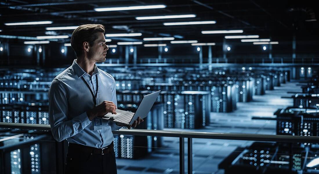 A man standing with a laptop in front of rows of servers.