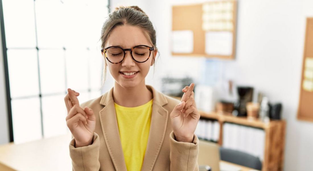 A young woman in business attire stands with her hands raised and fingers crossed in an office setting.