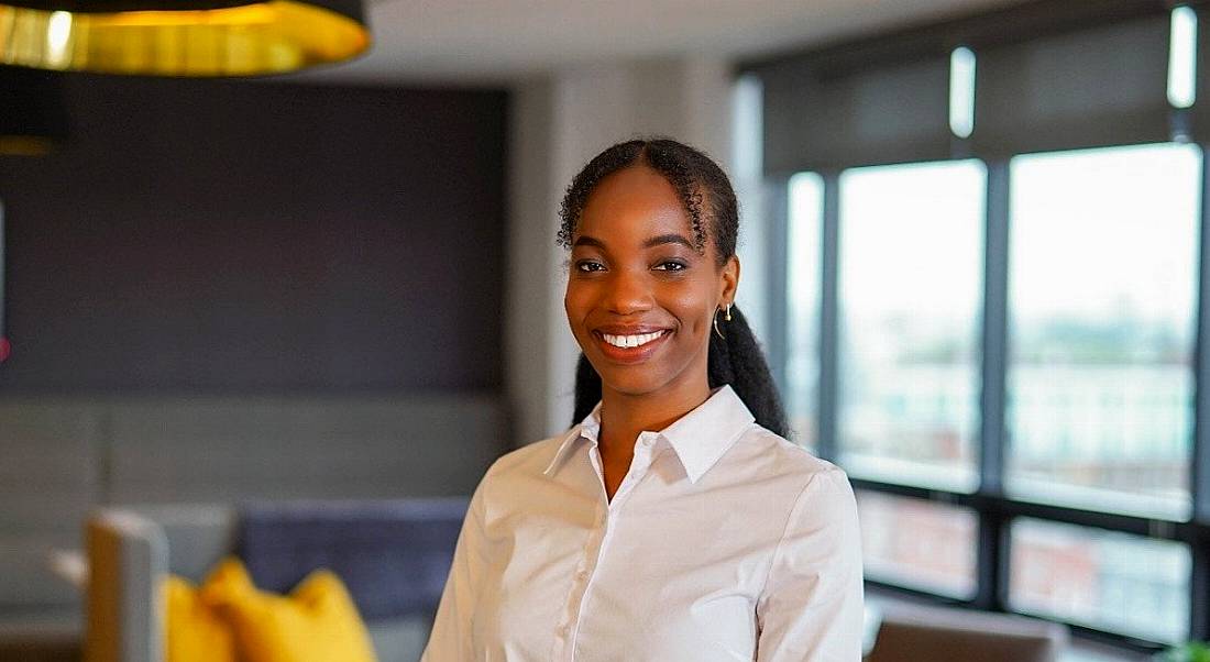 A young woman, who is a graduate, in business attire smiling at the camera in a well-lit room. There are grey couches and chairs in the background.