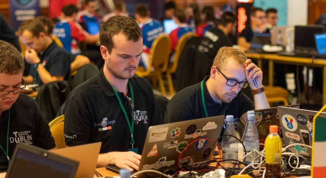 Three young men wearing black T-shirts working on computers in a busy hall. They are participating in a cybersecurity challenge.