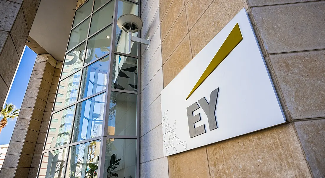 An EY logo on a white background on the side of a large office building on a sunny day.