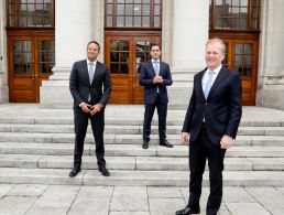 Three men stand outside Government buildings. In the centre is Leo Varadkar, holding up a tablet with the Money Jar website open on the screen.