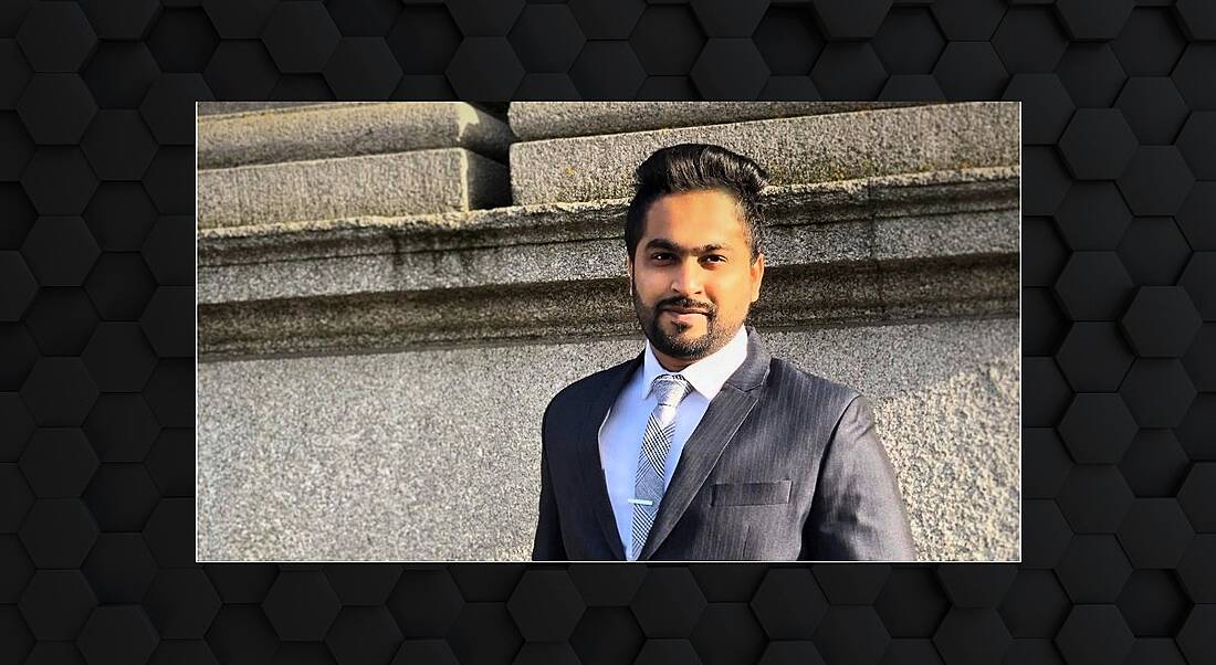 A young man in a business suit stands outside on a sunny day smiling at the camera. It is surrounded by a grey frame.