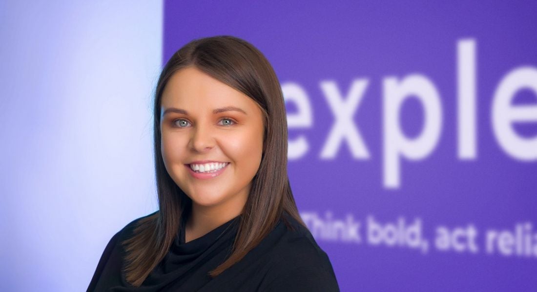 A young brunette woman smiling at the camera in front of a purple and white wall that says Expleo.