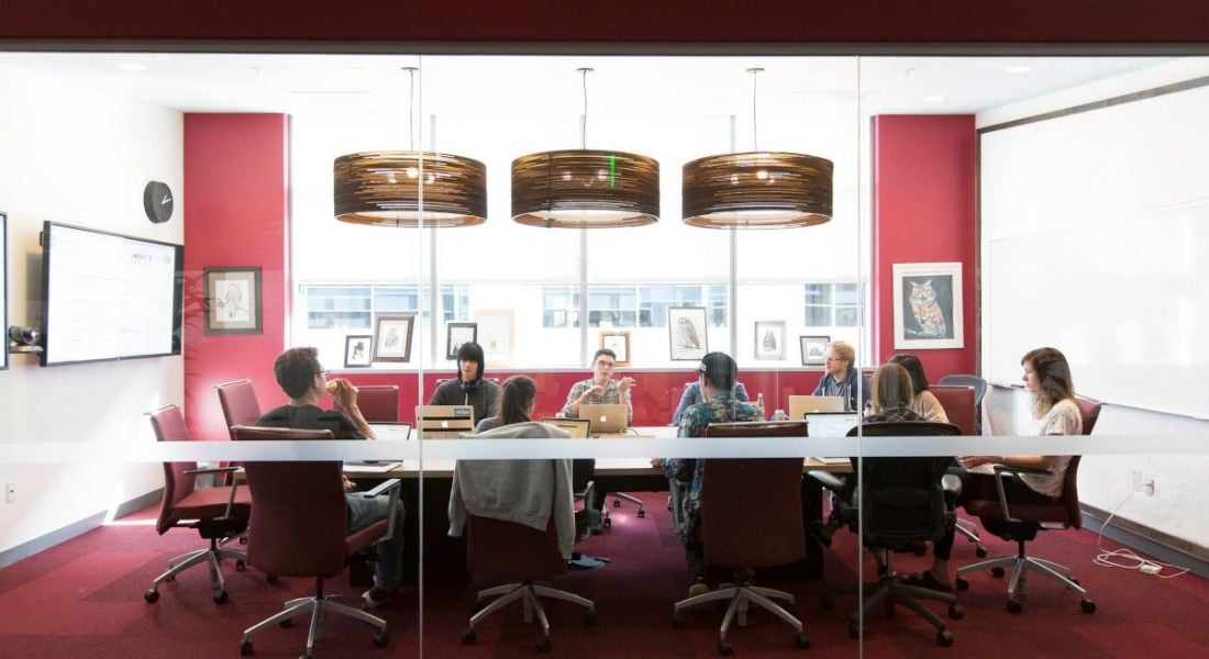 View through a glass-walled conference room with red carpet and red walls. 10 people are gathered around a large conference table.