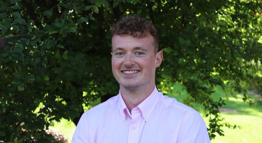 A young man wearing a pale pink shirt smiles at the camera against a backdrop of trees and foliage. He is a graduate working at Accenture.