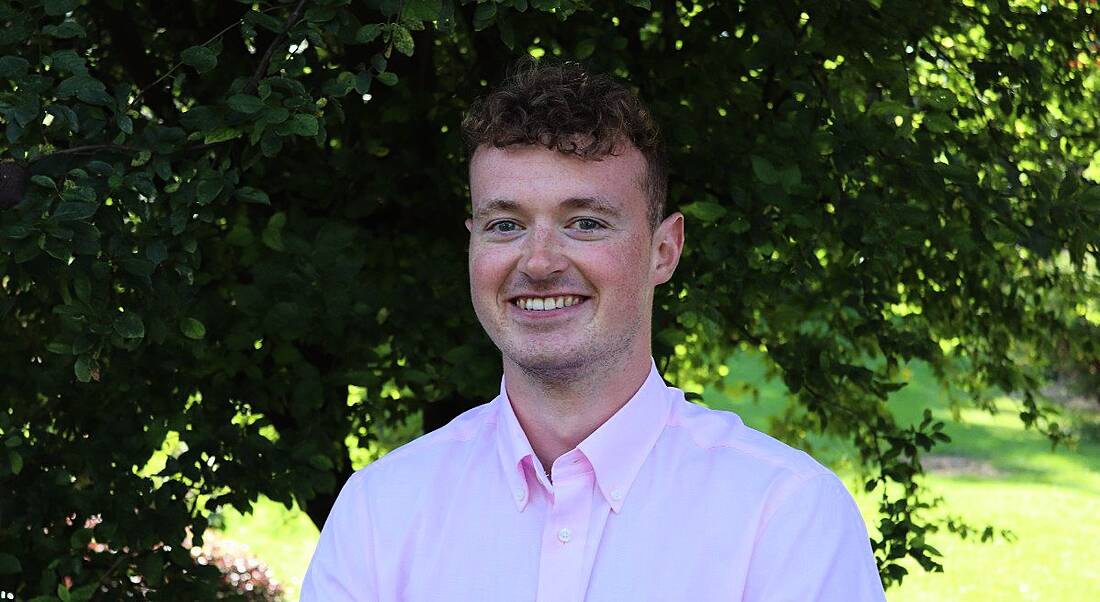 A young man wearing a pale pink shirt smiles at the camera against a backdrop of trees and foliage. He is a graduate working at Accenture.