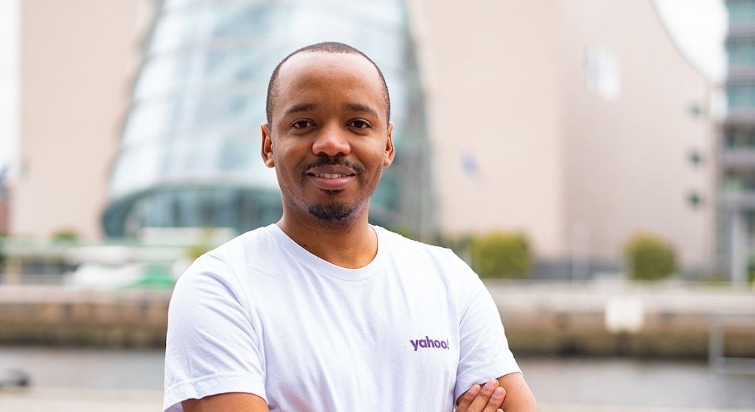A young man in a white T-shirt with the Yahoo logo on the left breast. He’s smiling at the camera with a cityscape in the background.
