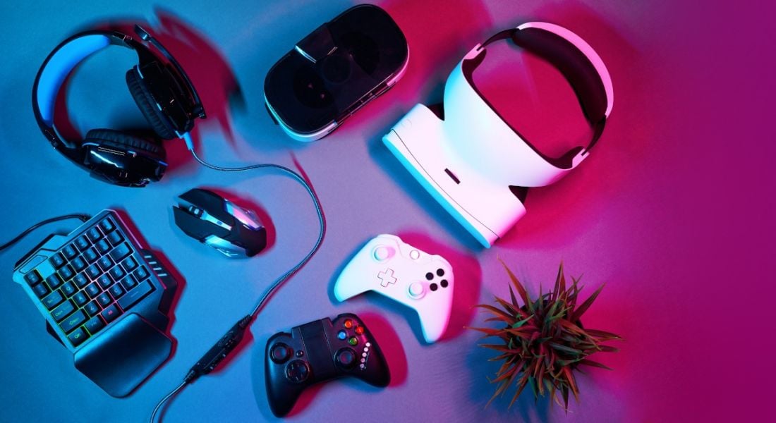Several gaming devices including controllers, headphones and a VR headset, sitting on a blue and pink neon-lit table.