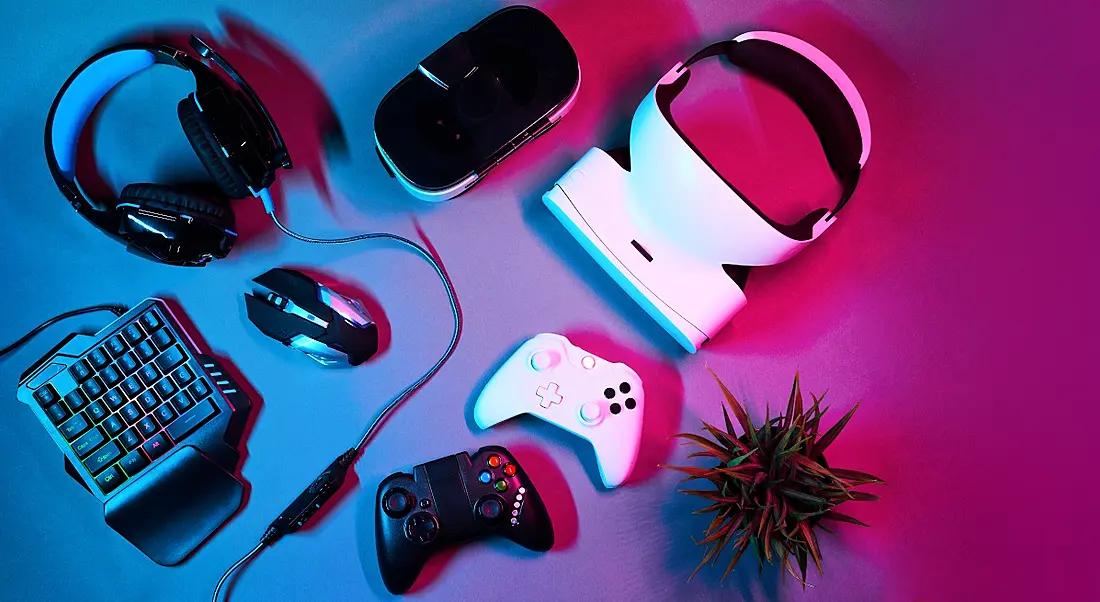 Several gaming devices including controllers, headphones and a VR headset, sitting on a blue and pink neon-lit table.