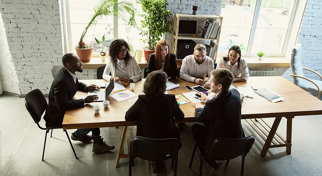 A group of Gen Z workers sit around a large table with an assortment of devices and notes, having a brainstorming session.