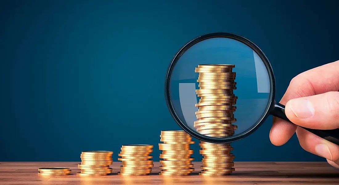 Stacks of coins are beside each other, gradually increasing in height. The final stack is the tallest and someone is holding a magnifying glass in front of the coins, indicating scrutiny over the gender pay gap.