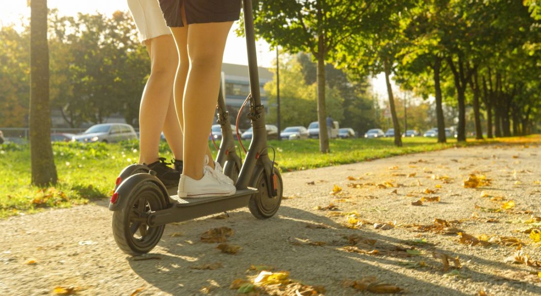 Two people can be seen riding on e-scooters. They are on a path and surrounded by trees. There is a warm glow from the sun which is casting a shadow in the direction of the photographer. Only the lower halves of the people are visible.