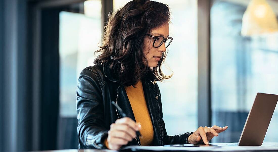 A woman with dark hair and glasses sits at desk working on a laptop, while taking notes beside it.