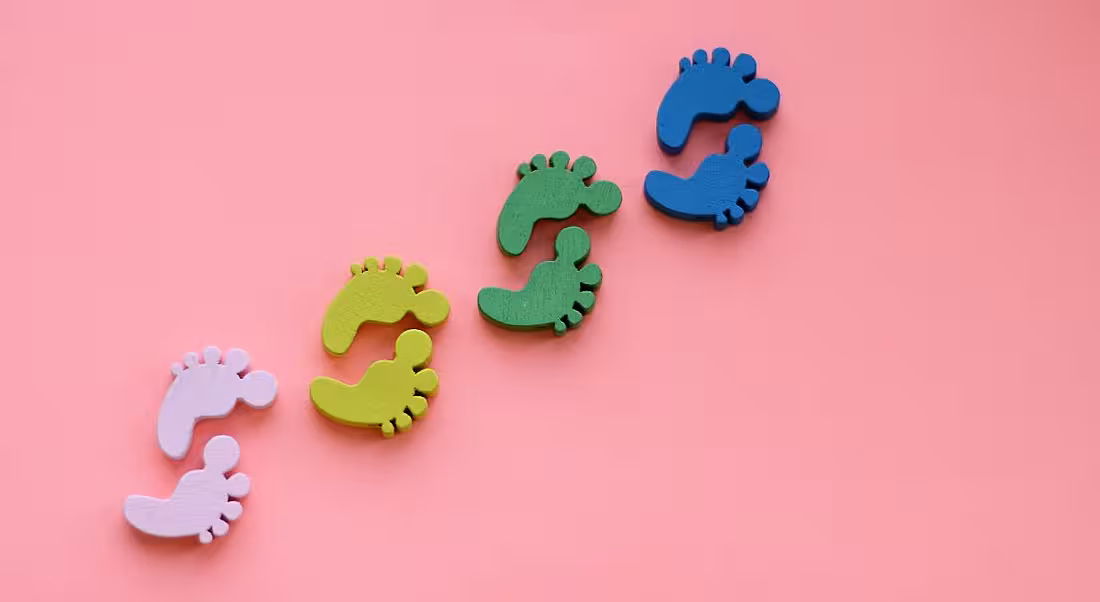 Pink, yellow, green and blue footprints against a pink background, symbolising first steps.