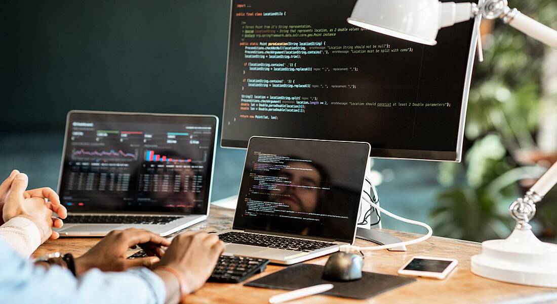 Two people are working on code at a desk with three screens and a white lamp.