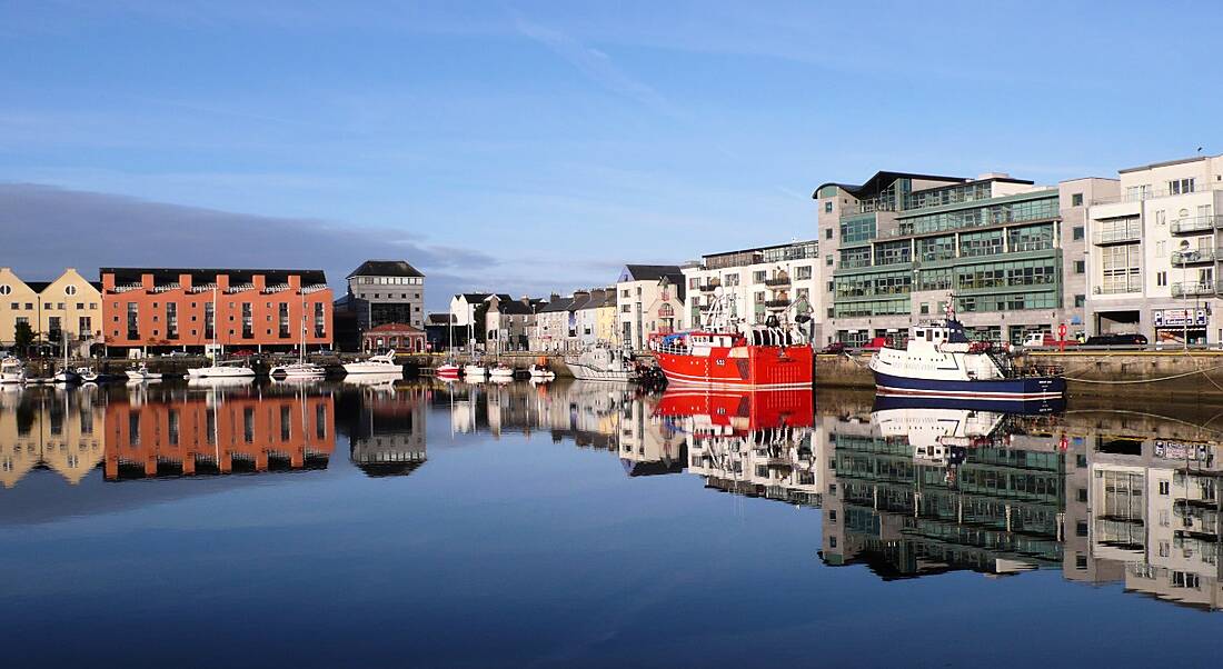 A still quay in Galway on a clear day, with boats docked, surrounded by buildings.