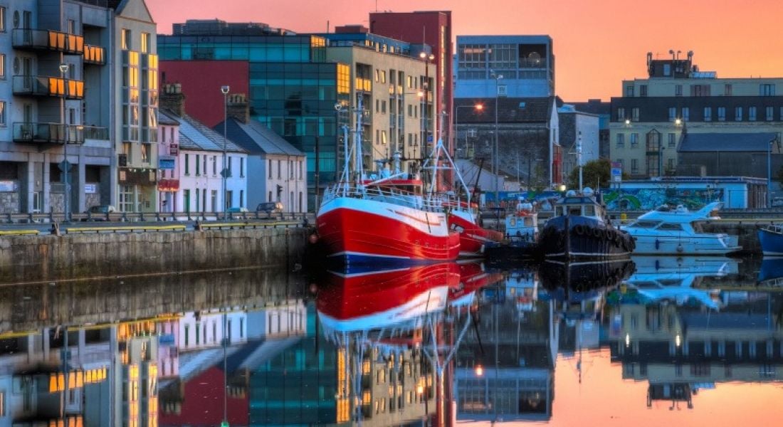 A sunrise over the docks in Co Galway. A red and white fishing boat features prominently against a row of buildings along the docks.