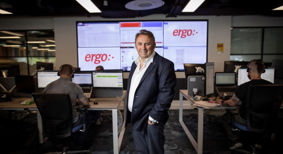 John Purdy stands in a suit in an office. Behind him is a screen that says 'Ergo'.