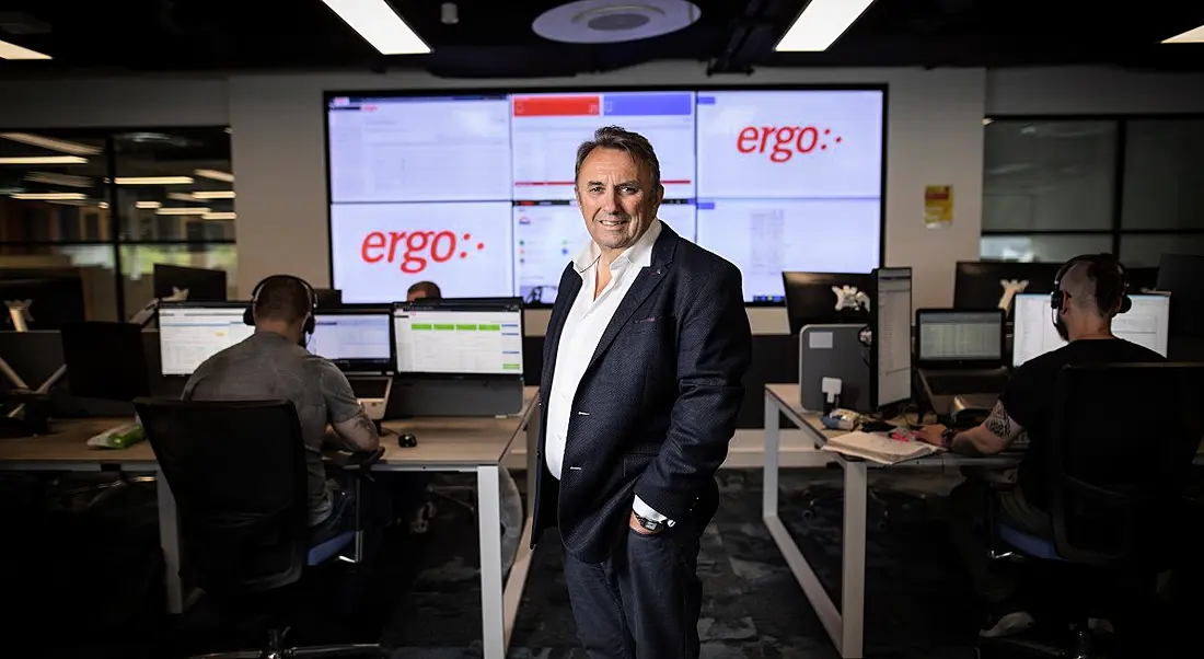 John Purdy stands in a suit in an office. Behind him is a screen that says 'Ergo'.