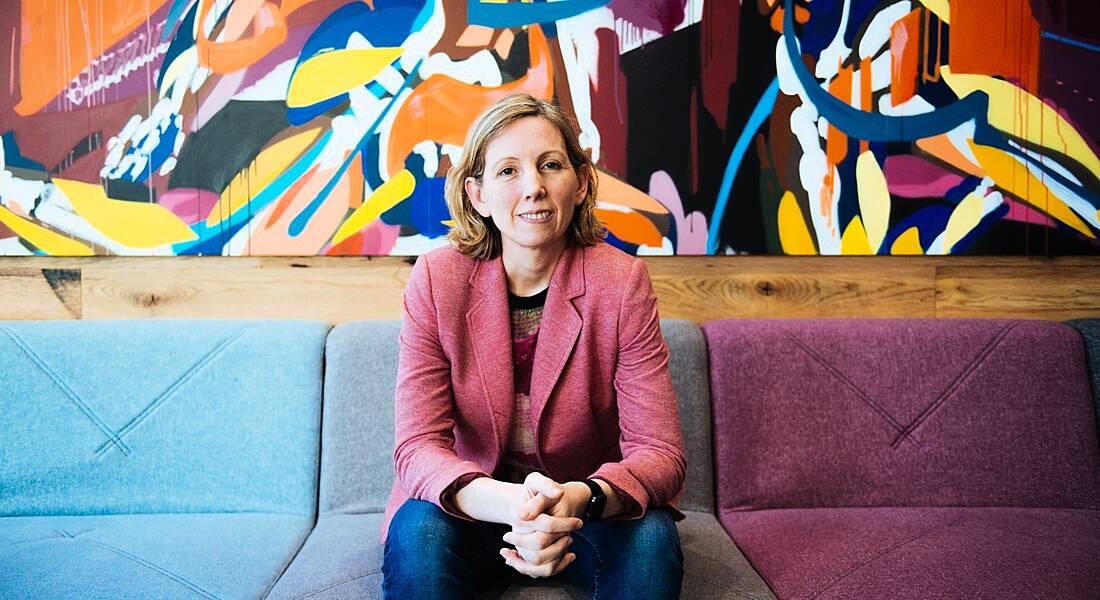 Medb Corcoran of Accenture Labs is sitting on a colourful couch in a colourful room, smiling into the camera.