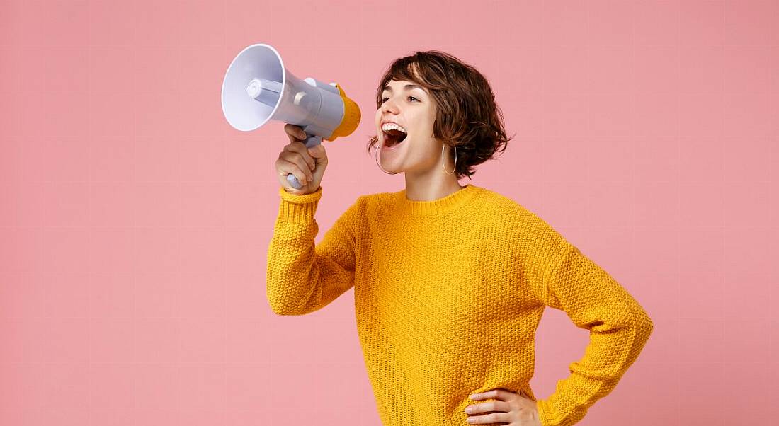 A woman with short hair and a yellow jumper is shouting into a megaphone.