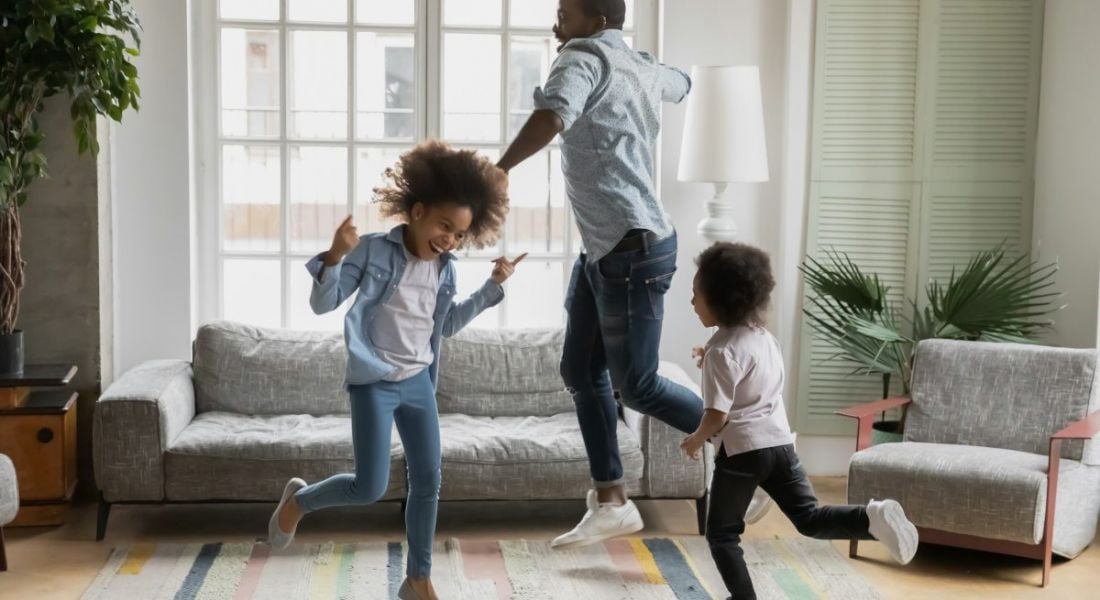 A father is getting his two children to move more at home, dancing and laughing in a bright living room.