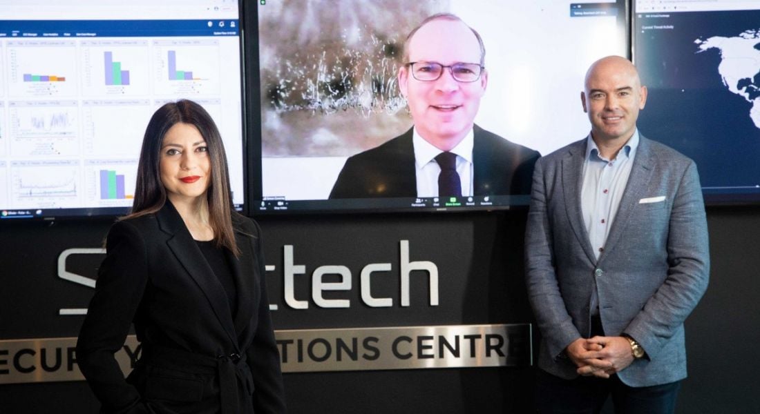 Raluca Saceanu and Ronan Murphy stand in front of a Smarttech247 logo with a large screen in between them. Minister Simon Coveney is on the screen.