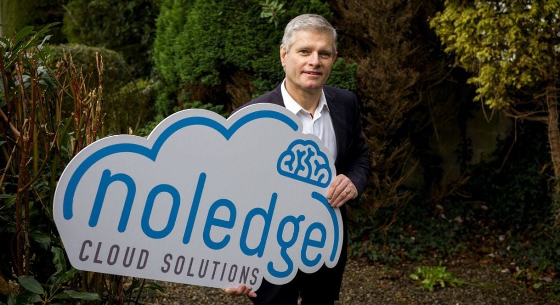 Ray Ryan, managing director of The Noeldge Group stands outside wearing a suit and holding a sign with the company's logo.