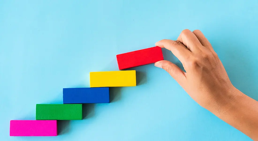 A hand places a red rectangular block on top of a bunch of other colourful blocks in the shape of a staircase against a blue background.