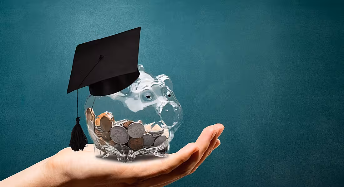A hand is holding up a clear piggy bank with a graduation hat on it against a green background.