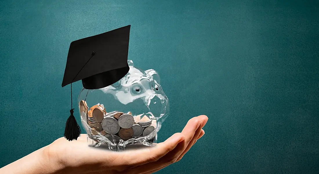 A hand is holding up a clear piggy bank with a graduation hat on it against a green background.