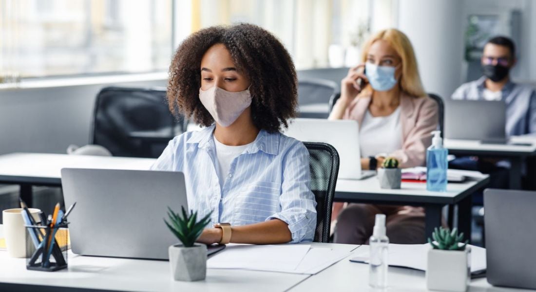 A group of young employees are working in an office, socially distanced and wearing masks.