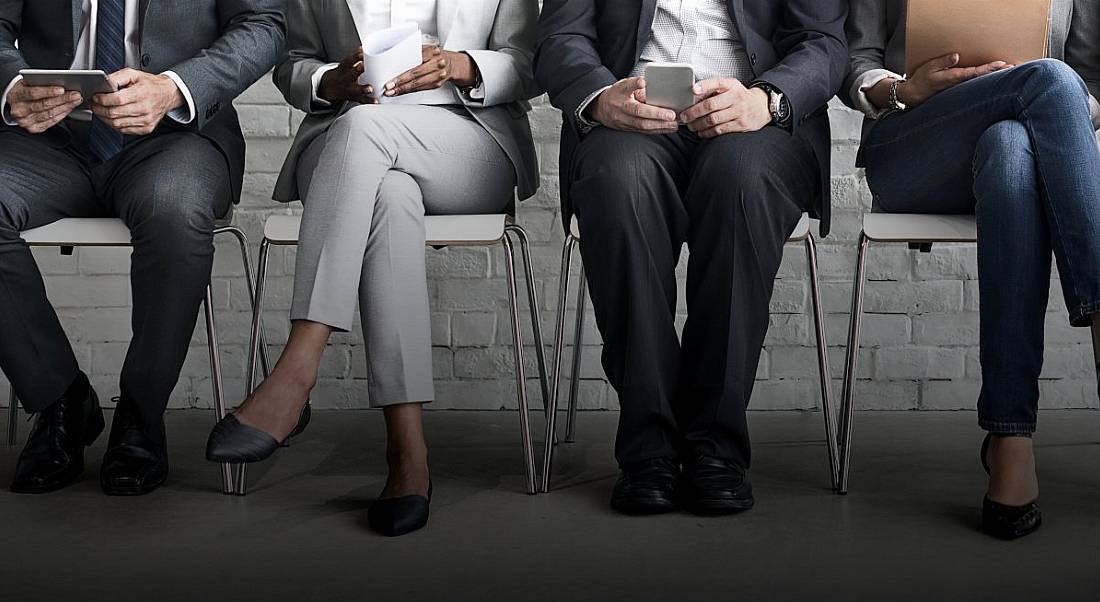 A group of job candidates are sitting side by side in a row, with just their legs and feet in frame.