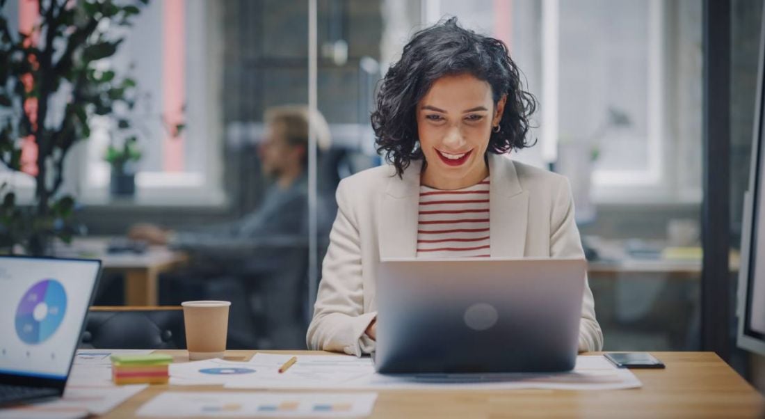 A woman is working at a laptop in a bright office space and smiling.