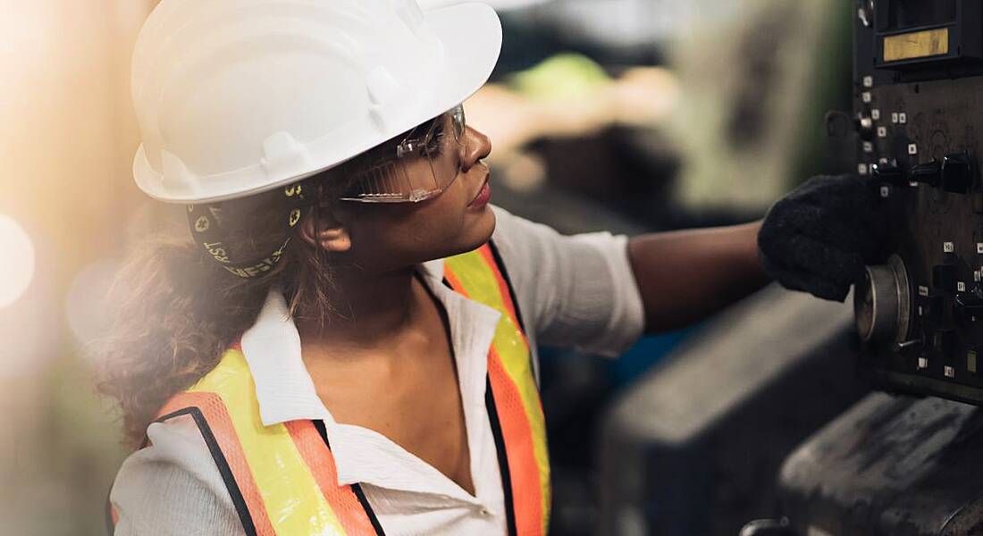 A woman in a hard hat, safety goggles and a high-vis jacket examining a piece of machinery.