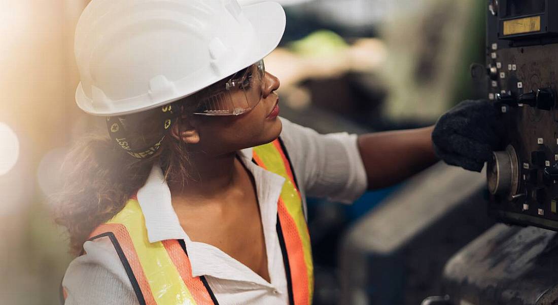 A woman in a hard hat, safety goggles and a high-vis jacket examining a piece of machinery.