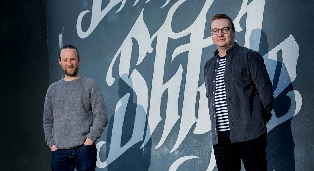 Two men from Frankli are standing in casual clothes in front of a wall with graffiti artwork on it.