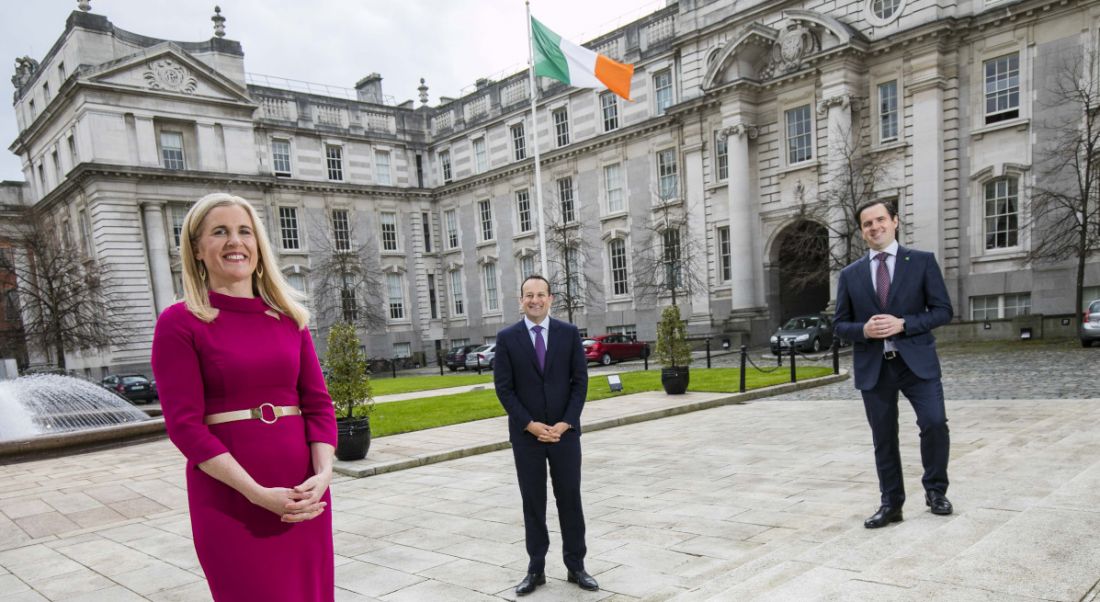 A woman and two men stand two metres apart in front of Government buildings bearing an Irish flag.