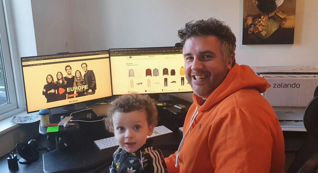 Jonathan Keenan of Zalando is working from home with his child sitting on his lap. He is wearing an orange hoodie and smiling into the camera while sitting at his desk.