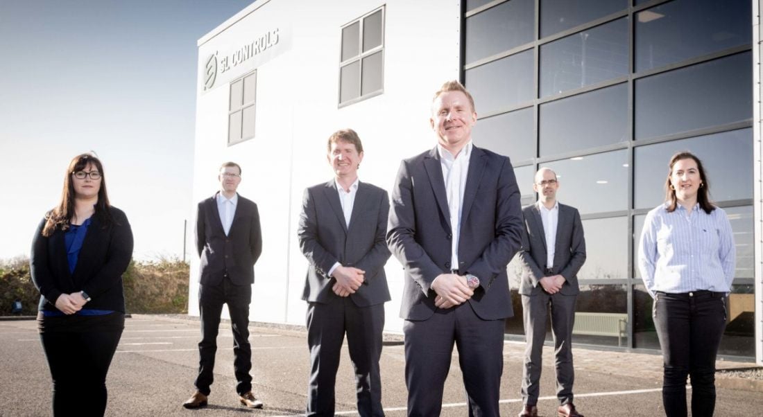 Six people in business attire stand outside in front of a large office building with the SL Controls logo on the side.