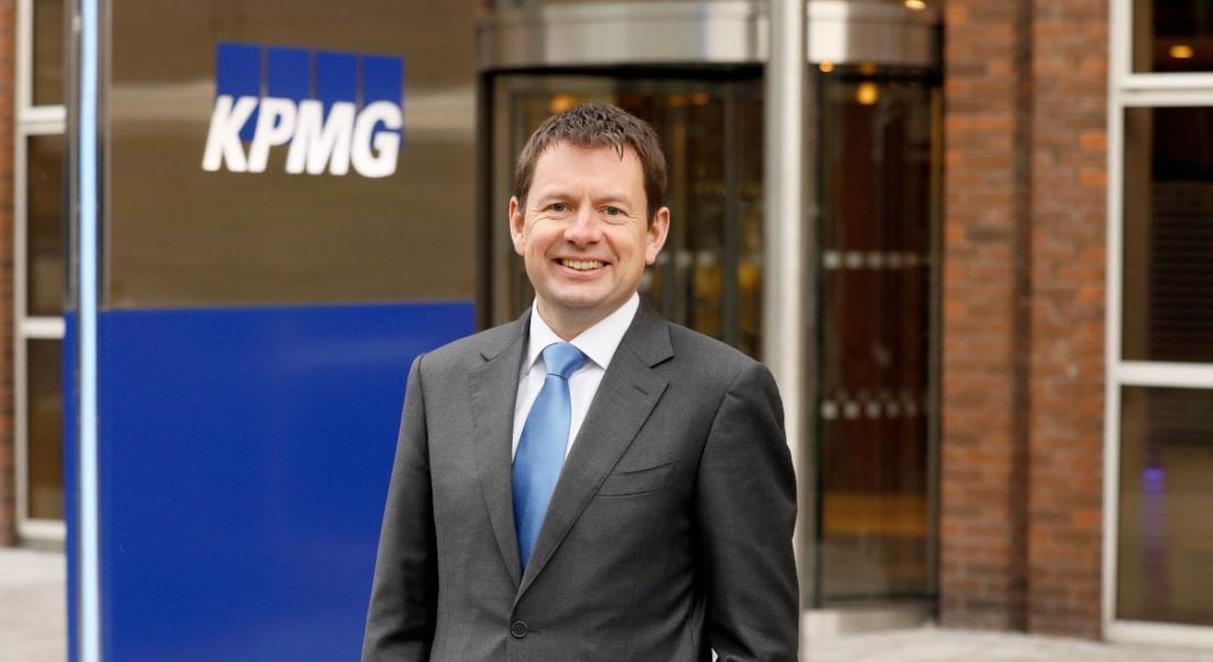 Seamus Hand stands in a suit outside an office building. The KPMG logo is behind him.