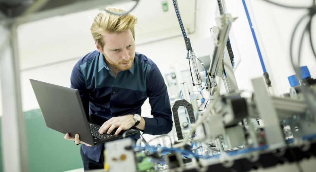 A male engineer holding a laptop while examining equipment in a factory.