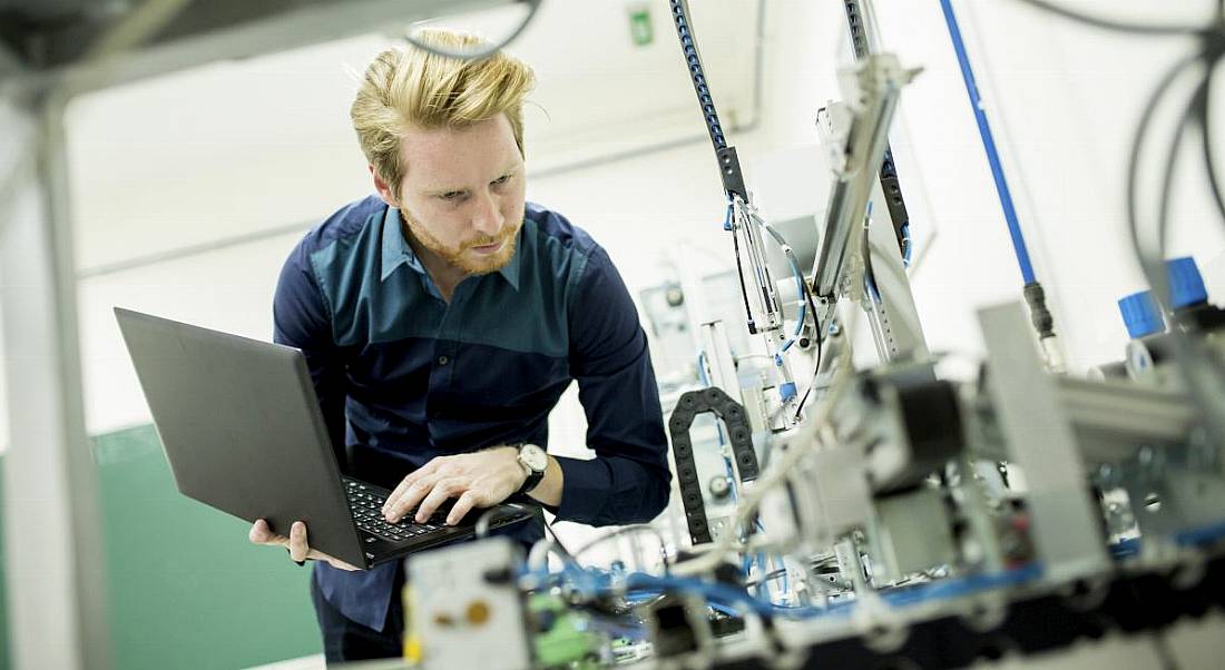 A male engineer holding a laptop while examining equipment in a factory.