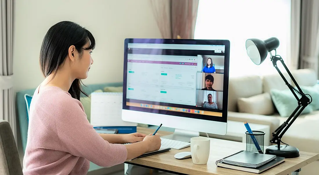 A woman remote working at home at a desk with a large computer monitor. She’s taking notes while attending a virtual meeting.