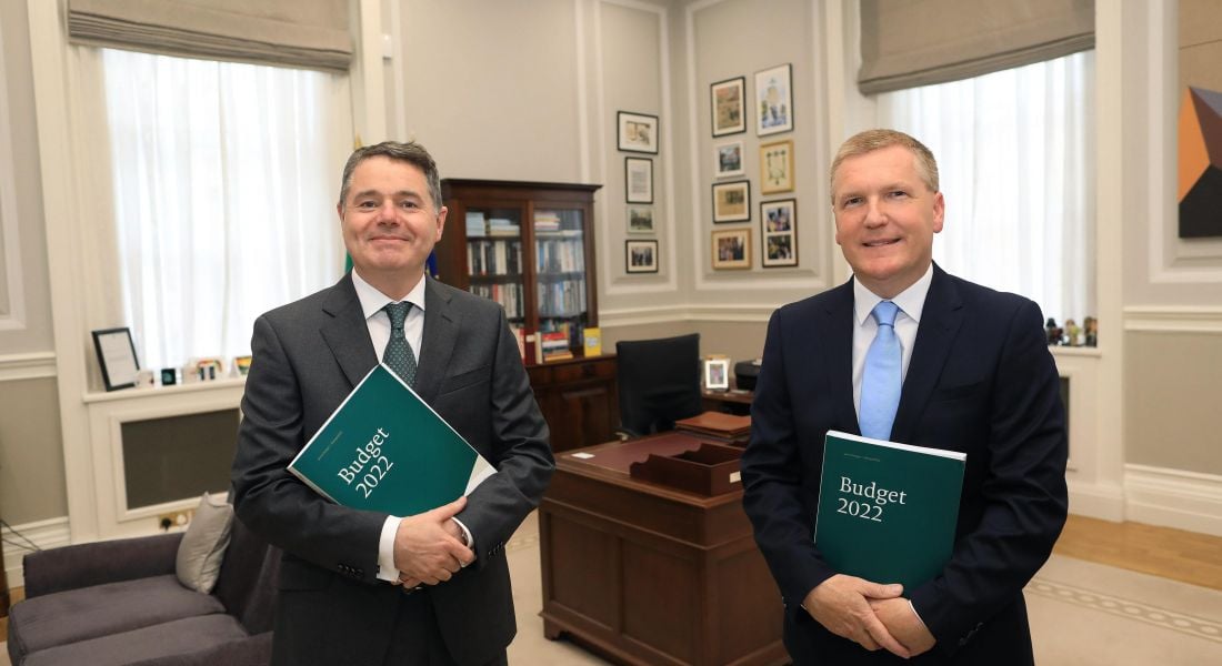 Two men in suits stand in a Government office smiling at the camera. Both are holding green folders that say Budget 2022 on the front.