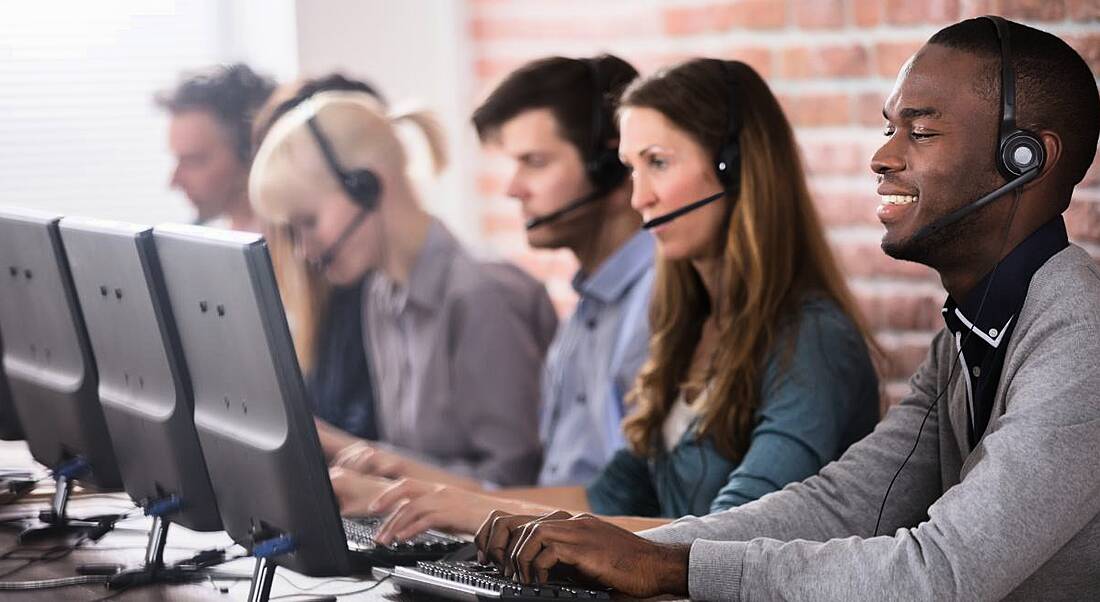 Five office workers sitting in a row at computers with headsets on the phone to customers.