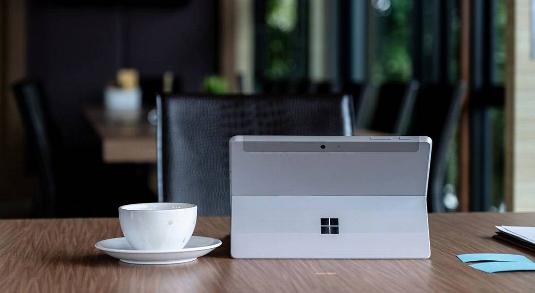 Microsoft logo on a silver tablet sitting on a desk with a coffee cup and paper notes.